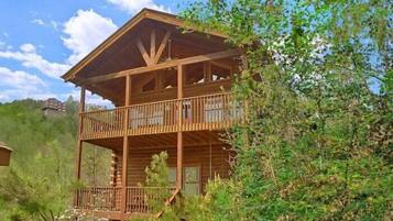 A two-story wooden cabin with a balcony, surrounded by greenery.