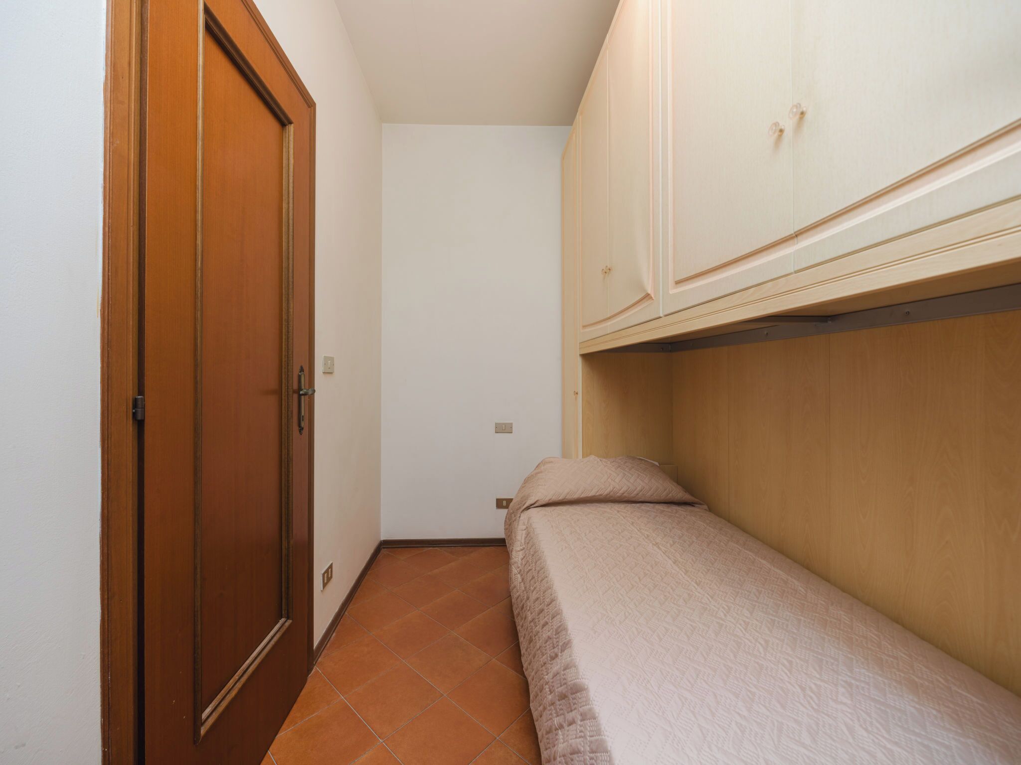 A narrow room with a wooden door, a bed with a headboard, and a white wardrobe.