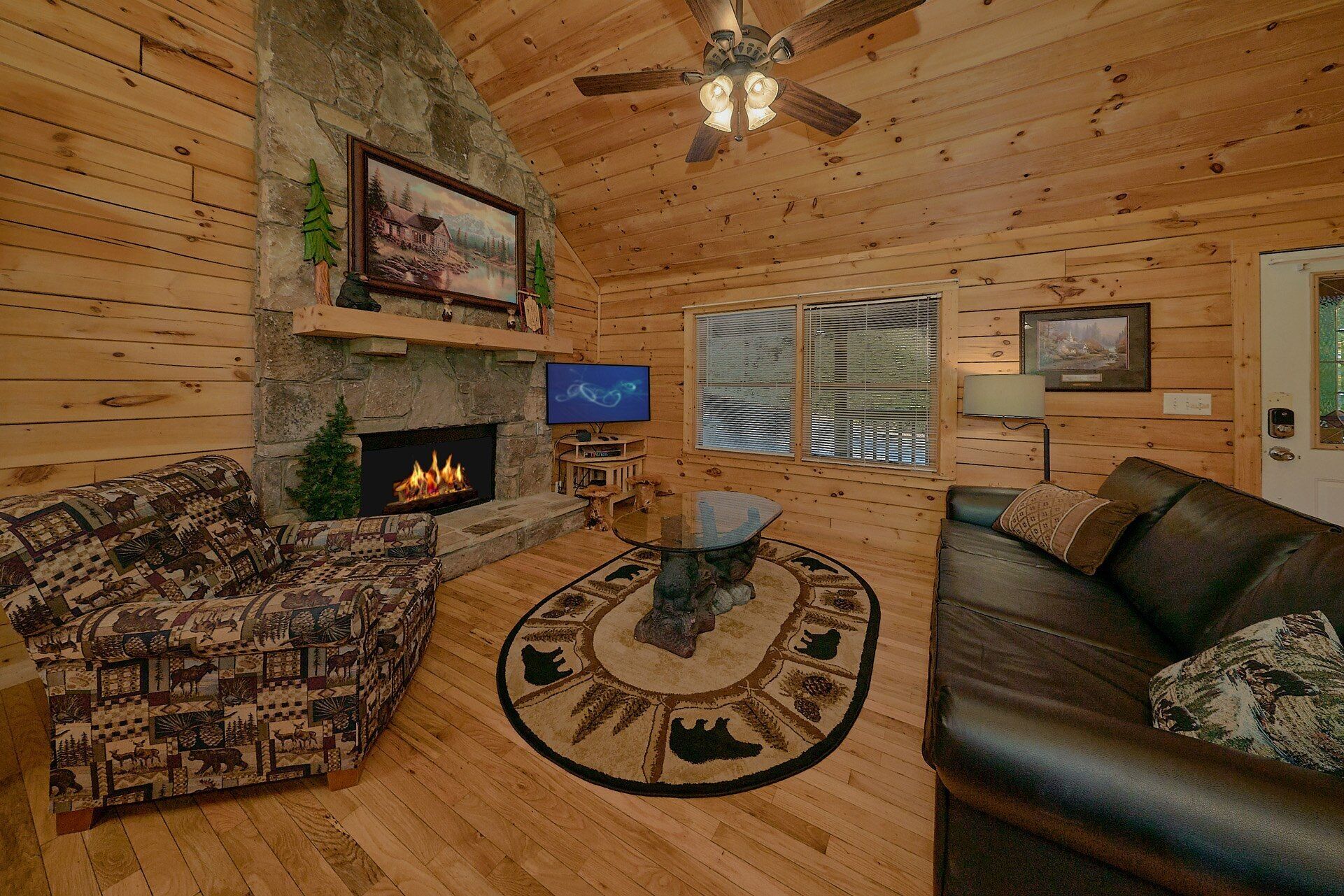 A log cabin interior with a fireplace, a television, a ceiling fan, and a large window.