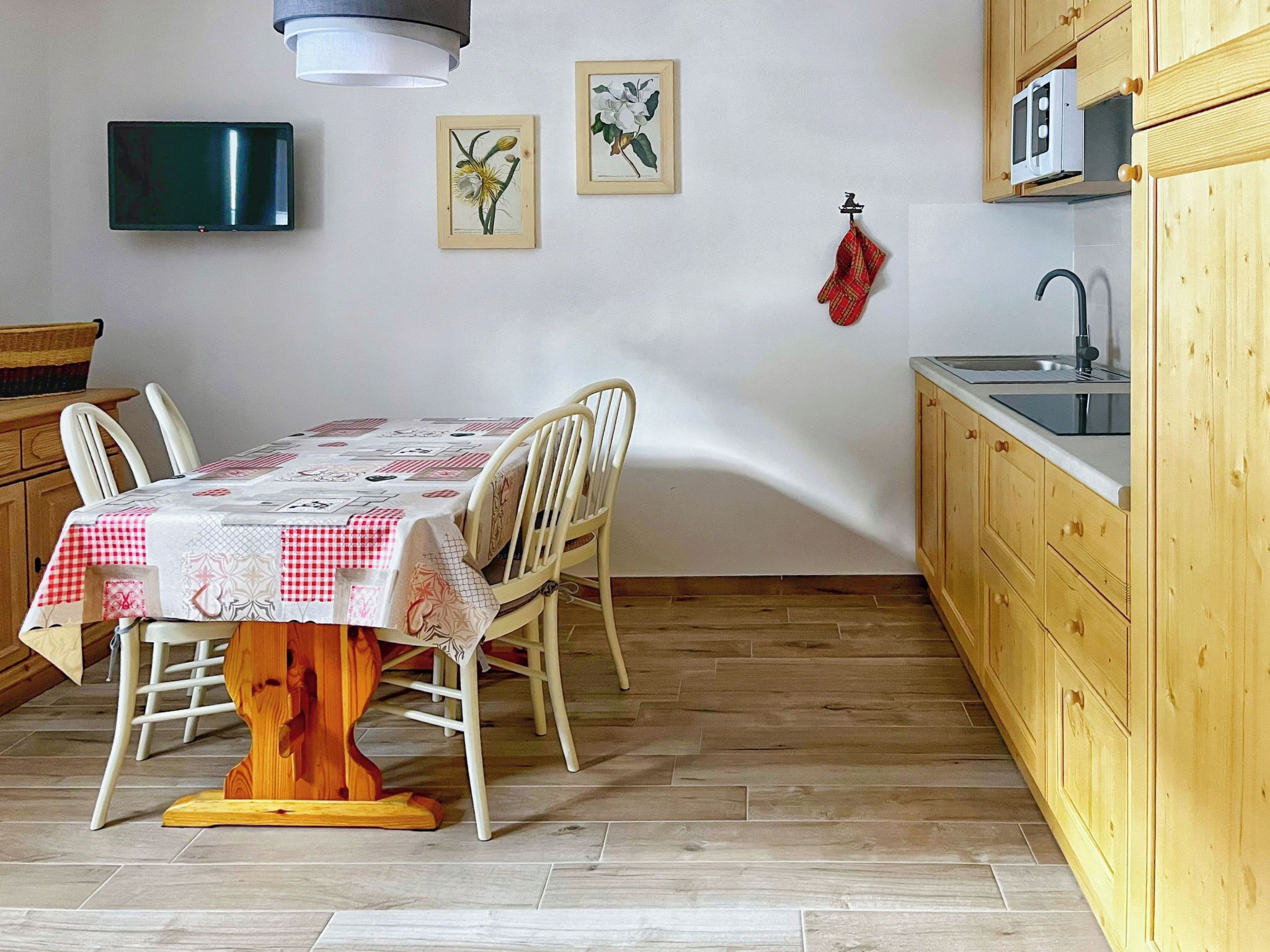 A dining area with a table set for four, wooden chairs, a wooden dining table, a kitchen with wooden cabinets, a microwave, and a sink.