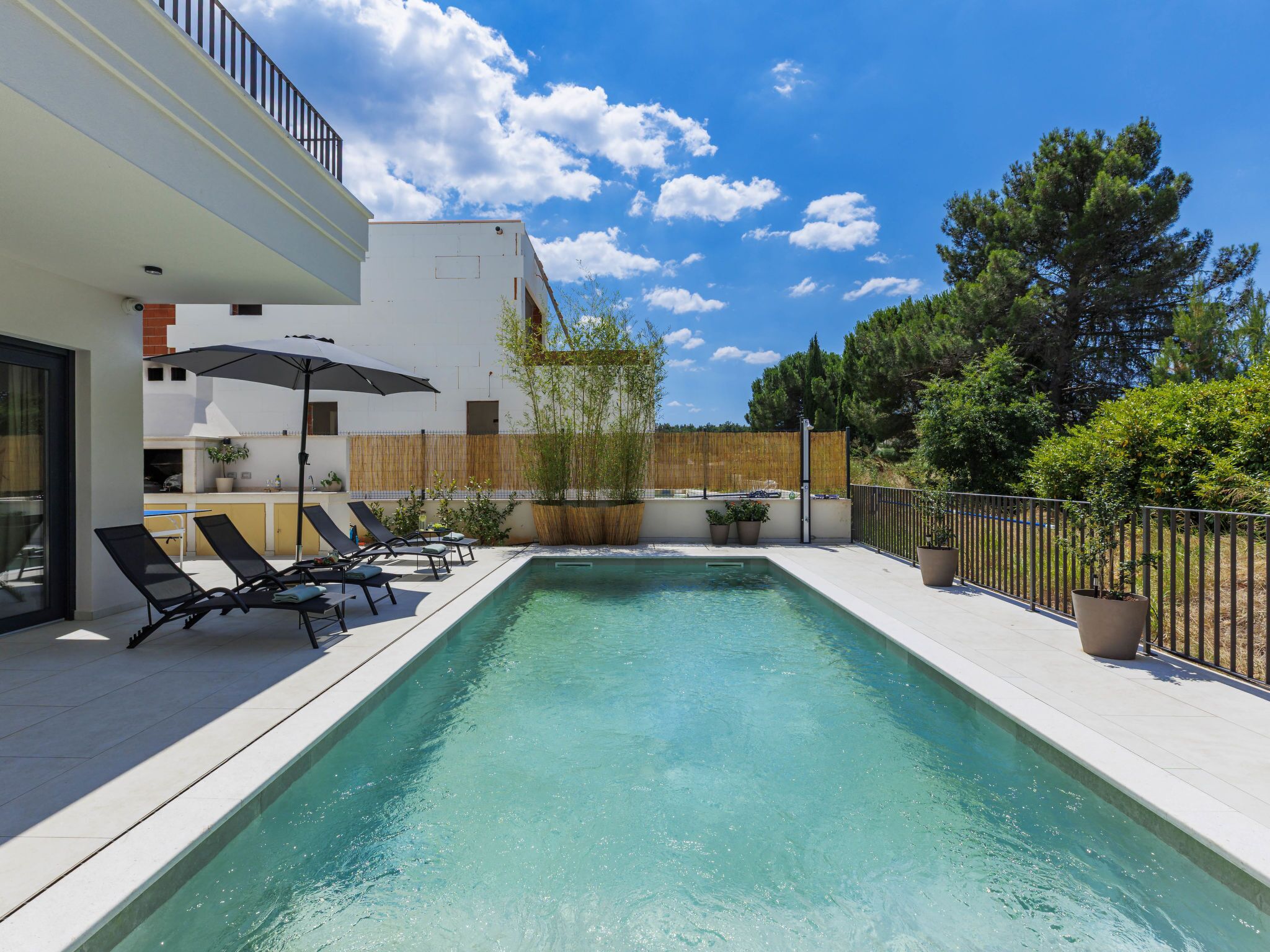 A rectangular swimming pool with lounge chairs and umbrellas, surrounded by a wooden fence and trees.