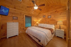 A wooden-paneled bedroom with a bed, bedside tables, a ceiling fan, and a television mounted on the wall. - A Mountain Lodge | Hot Tub | Game Room (Pigeon Forge)
