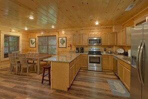 A wooden cabin-style kitchen with a central island, wooden cabinets, stainless steel appliances, and a dining area with chairs. - A Mountain Palace | Theater Room | Hot Tub (Pigeon Forge)