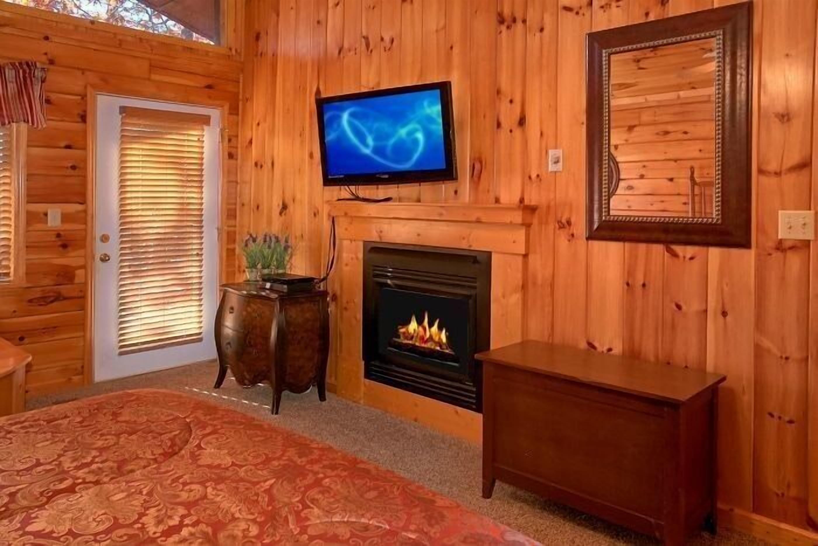 Una habitación con paneles de madera, un televisor sobre la chimenea, una cama con colcha estampada roja y una pequeña mesa de madera.