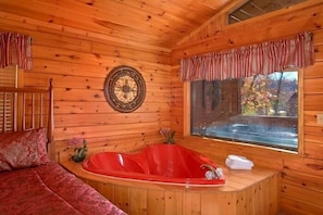 A wooden cabin interior with a red bathtub, a wooden bed, and a window with a view of trees. - A New Beginning | Hot Tub | Close to Dollywood (Sevierville)