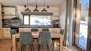 A modern kitchen with a large dining table, wooden cabinets, and a view of a snowy landscape. - Rebelka by Interhome (Horni Misecky)