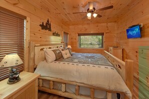 Camera da letto in una baita di legno, con un letto, un comodino, una lampada, una ventola a soffitto, una finestra e un televisore.
