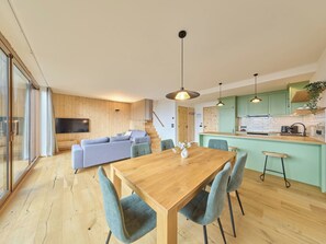 Un espai de vida modern amb taula de dinar de fusta, sofà tapissat de gris i una zona de cuina amb armaris verds i il·luminació penjant.