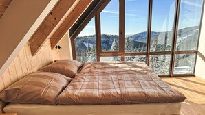 A bedroom with a wooden bed, large windows, and a view of snow-covered mountains. - Rebelka by Interhome (Horni Misecky)