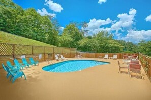 A swimming pool with lounge chairs and tables, surrounded by a wooden fence and greenery. - A Romantic Journey | Honeymoon Cabin with Hot Tub (Pigeon Forge)