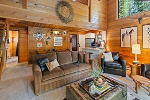 A cozy cabin interior with a wooden ceiling, a sofa, a coffee table with books and decorative items, and a kitchen area in the background. - Trail One | Black Butte Ranch | Sleeps 8 | Family (Sisters)