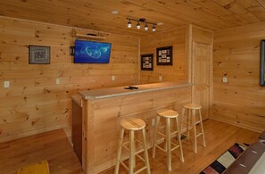 A wooden cabin interior with a bar area, stools, a television, and framed pictures on the walls. - A Spectacular View to Remember | Mountain Views | Hot Tub (Gatlinburg)