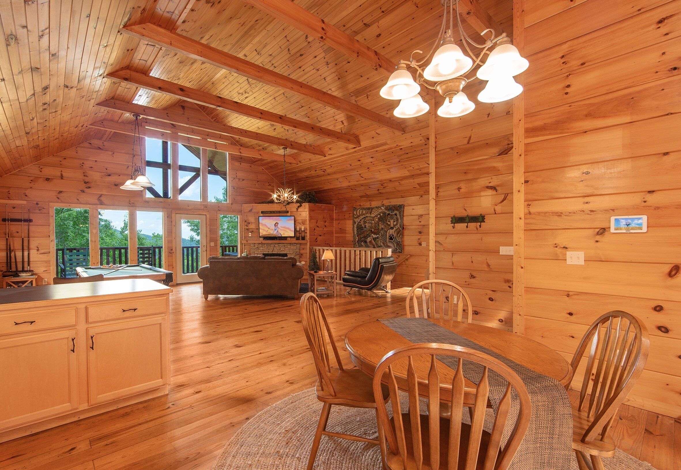 A wooden cabin interior with a dining area, kitchen, and a large window overlooking greenery.