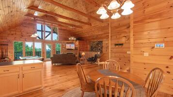 A wooden cabin interior with a dining area, kitchen, and a large window overlooking greenery.