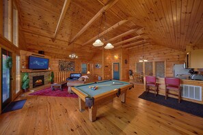 A wooden cabin interior with a pool table, seating area, fireplace, and a television.