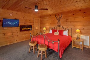A wooden cabin bedroom with a bed, two stools, a television, and a fireplace.