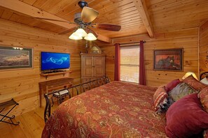 A wooden cabin interior with a bed, a television, a ceiling fan, and a window with blinds. - Angel's Landing Cabin Rental (Gatlinburg)
