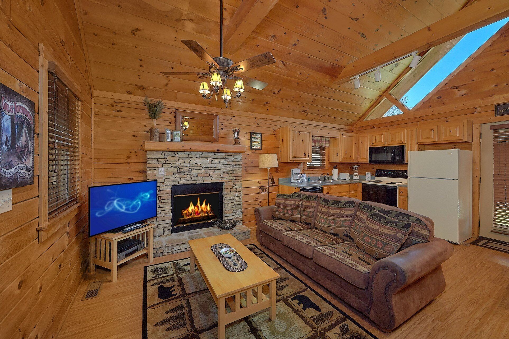 A cozy cabin interior with a stone fireplace, wooden furniture, a television, and a kitchen area.