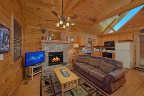 A cozy cabin interior with a stone fireplace, wooden furniture, a television, and a kitchen area. - Angel's Ridge Cabin Rental (Pigeon Forge)