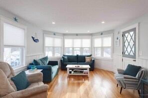 A living room with a blue sofa, a striped armchair, and a coffee table. There are multiple windows providing natural light. - Beautiful ocean view home in Old Orchard Beach! (Old Orchard Beach)