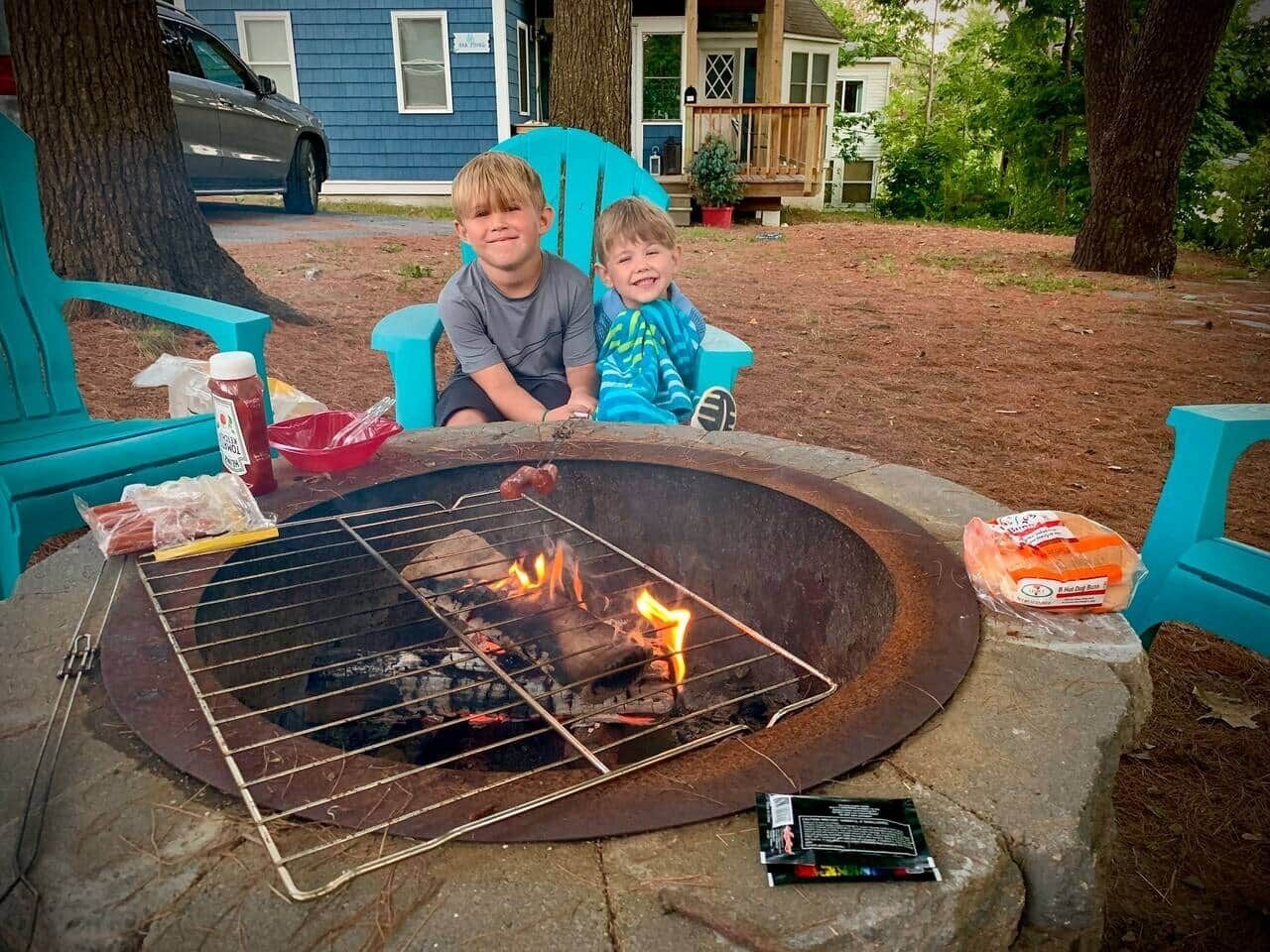 Two children sitting at a campfire with a grill, surrounded by outdoor furniture and a house in the background.