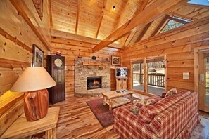 A log cabin interior with a fireplace, wooden walls, a sofa, and a table. - Apache Sunset Cabin Rental (Wears Valley)