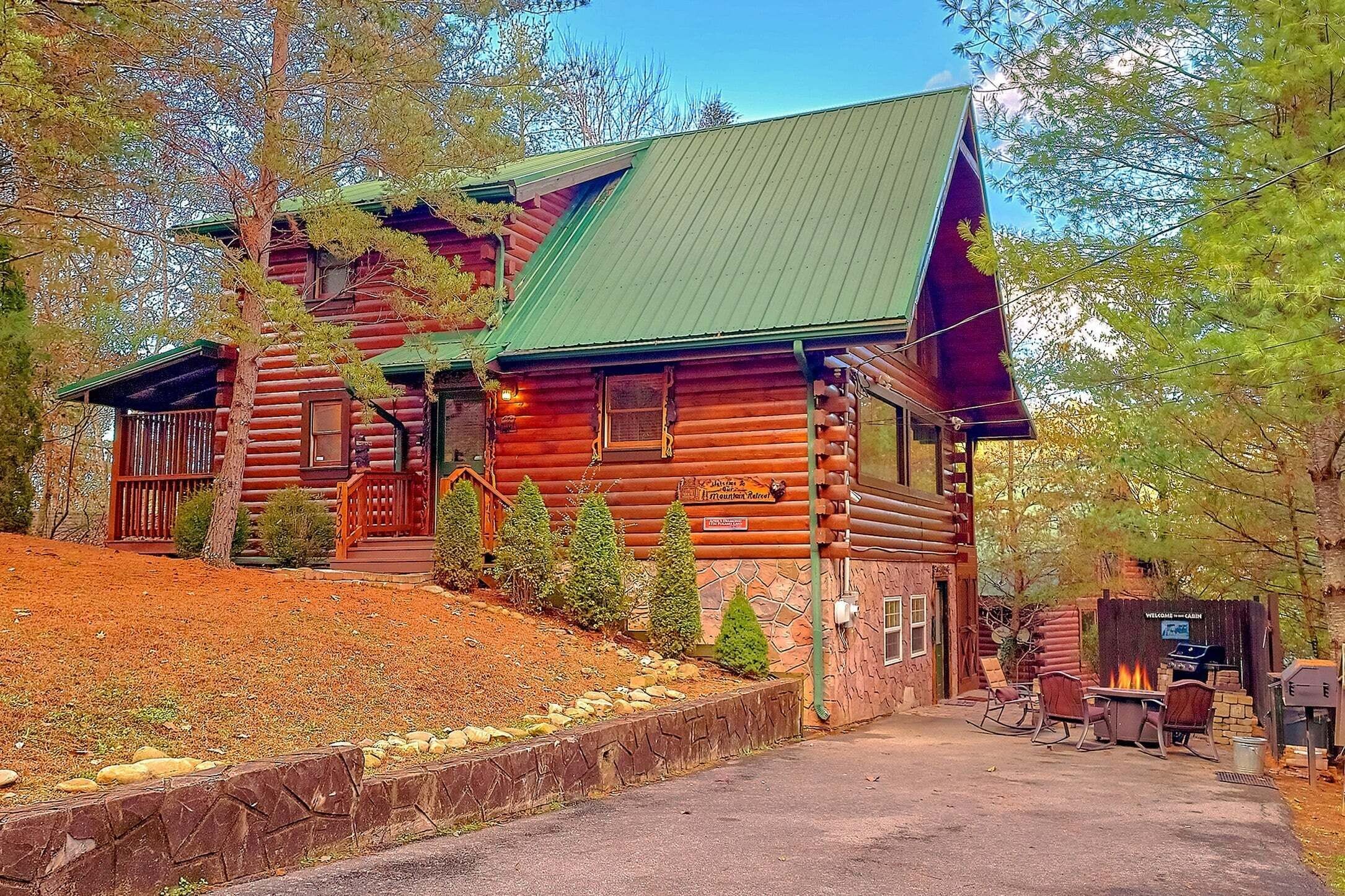 A log cabin with a green roof, a stone foundation, and a fire pit area with seating.