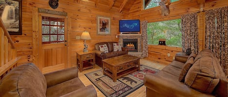 A cozy log cabin interior with a fireplace, leather sofas, a television, and a wooden ceiling.