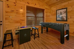 A wooden room with a foosball table, a bar area with stools, and a window with blinds. - Bear Cove Lodge Cabin Rental (Pigeon Forge)