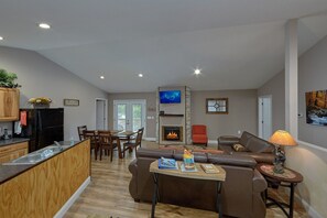 A modern living room with a fireplace, a dining area, and a kitchen with a refrigerator. - Bearfoot Bungalow | Hot Tub | Close to Dollywood | Free Attraction Tickets (Pigeon Forge)