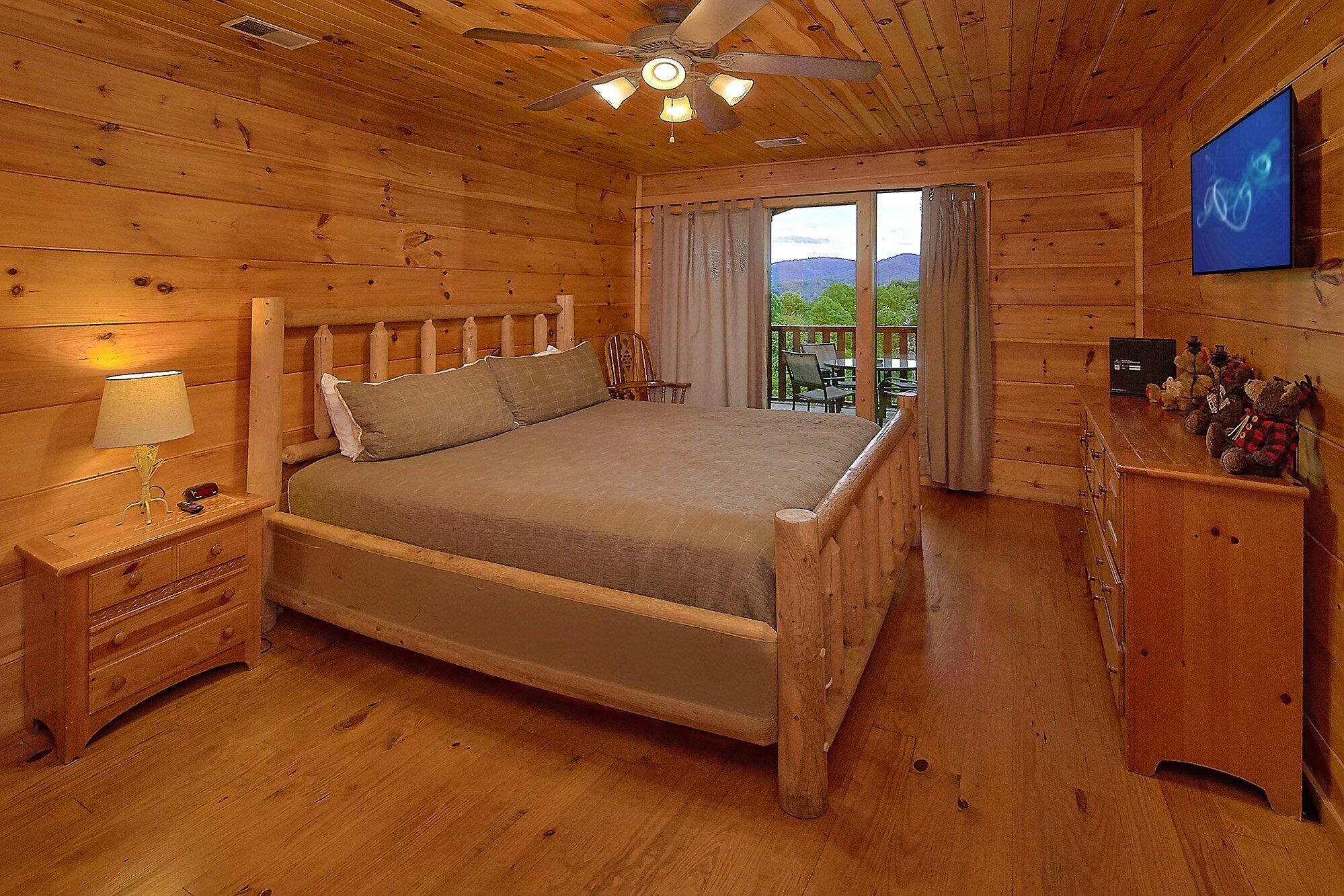 Een slaapkamer in een houten chalet met een bed, nachtkastje, lamp, plafondventilator en uitzicht op de natuur.