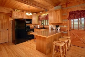 A wooden cabin interior with a kitchen area, including a refrigerator, stove, and bar stools. - Bearway to Heaven Cabin Rental (Wears Valley)