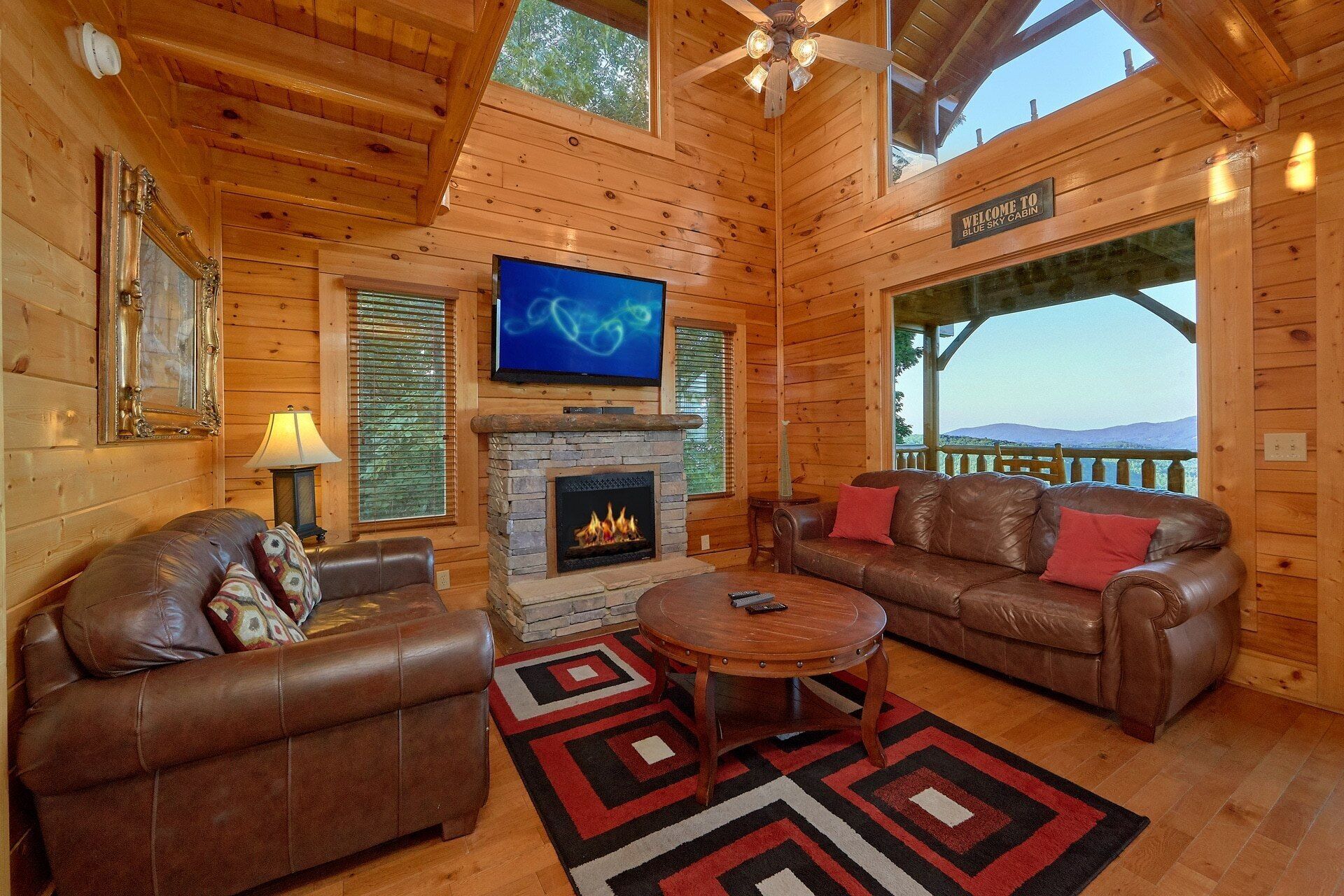 A log cabin interior with a fireplace, leather sofas, a television, and a scenic view.
