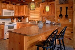 A wooden cabin interior with a kitchen featuring wooden cabinets, a central island with bar stools, a microwave, a stove, and a hanging light fixture. - Catch of the Day | Mtn Views, Hot Tub, Game Room, Pigeon Forge, Fireplace + Free Attraction Tickets (Pigeon Forge)