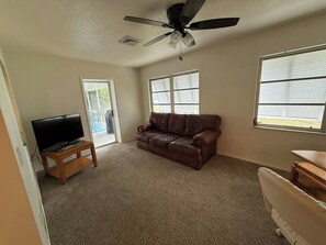 Una sala de estar con un sofá de cuero marrón, un mueble bajo de madera para la televisión, un ventilador de techo y una ventana con persianas.