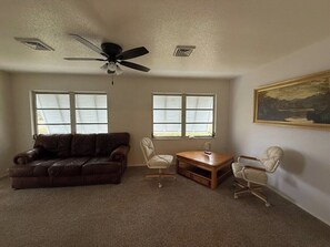 A living room with a brown leather couch, two white chairs, a wooden coffee table, and a ceiling fan. - The Manasota Retreat - Steps from Lemon Bay Park, Private Lanai w/ BBQ (Englewood)