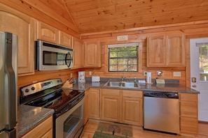 A kitchen with wooden cabinets, a microwave, a stove, a dishwasher, and a window. - Cheeky Chipmunk Getaway (Sevierville)