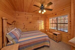 A wooden cabin bedroom with a bed, a nightstand, a window, and a ceiling fan. - Cheeky Chipmunk Getaway (Sevierville)