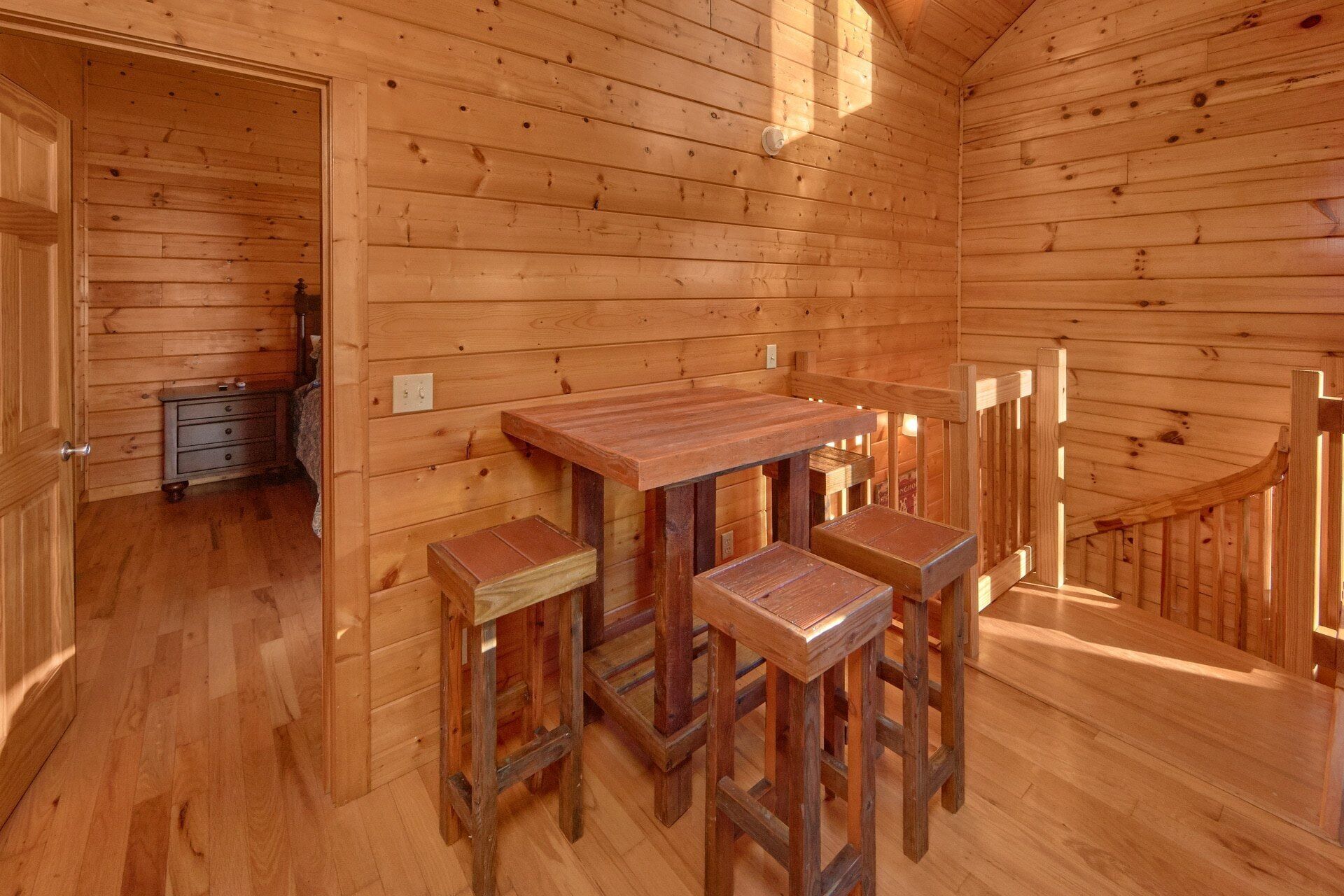 A wooden interior with a dining table and four stools, a staircase, and a door leading to another room.