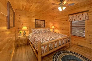 A wooden cabin bedroom with a bed, two bedside lamps, a ceiling fan, a window with blinds, and a framed picture on the wall.