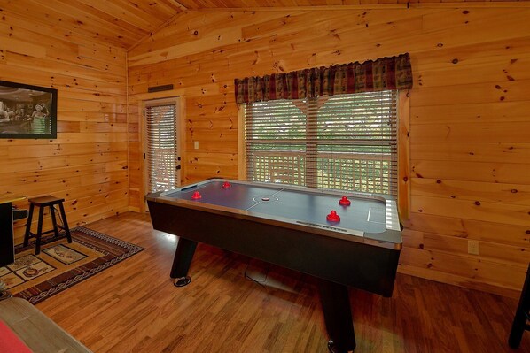A wooden-floored room with a foosball table, a window with blinds, and a framed picture on the wall.
