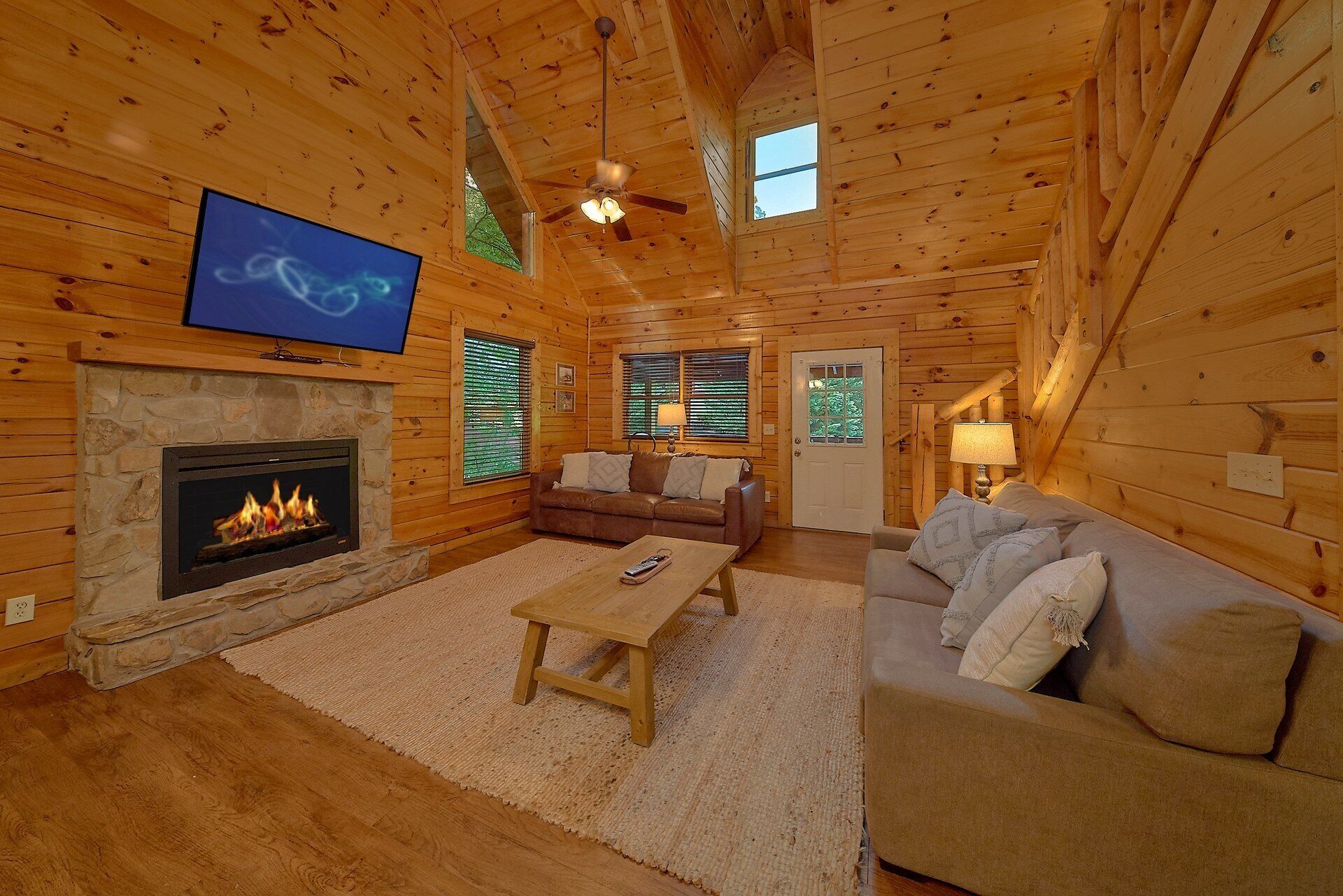 A log cabin interior with a stone fireplace, a television mounted on the wall, a ceiling fan, and a wooden floor.