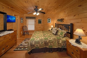 Ein Schlafzimmer in einer Holzhütte mit einem Bett, einem Sessel, einem Fernseher und einem Deckenventilator.