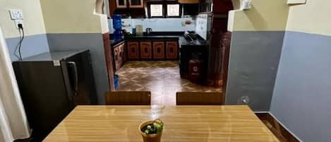 A dining area with a wooden table and chairs, a small potted plant, and a view into a kitchen with cabinets and a water dispenser.