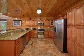 A kitchen with wooden cabinets, granite countertops, stainless steel appliances, and a stone tile floor. - Elk Ridge Lodge | Sleeps 22, Indoor Pool, Theater, Mtn Views, Hot Tub + Free Attraction Tickets (Pigeon Forge)