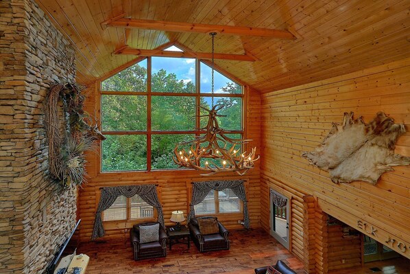 A log cabin interior with a large window, a chandelier, leather chairs, and a fur rug.