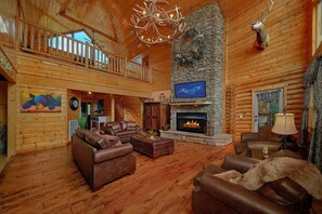 A spacious log cabin interior with a fireplace, leather sofas, a mounted deer head, and a chandelier.