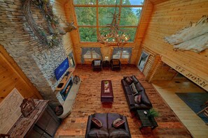 A log cabin interior with a stone fireplace, leather sofas, a chandelier, and a large window offering a view of trees.