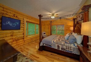 Une chambre située dans une cabane en rondins, avec un lit à baldaquin, un ventilateur de plafond et une grande fenêtre équipée de stores.