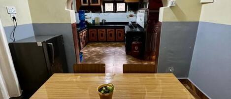 A dining area with a wooden table and chairs, a small potted plant, and a view into a kitchen with cabinets and a water dispenser.